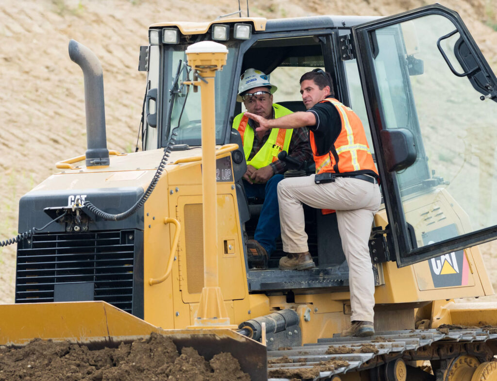 Two workers discussing in bulldozer cabin at construction site.